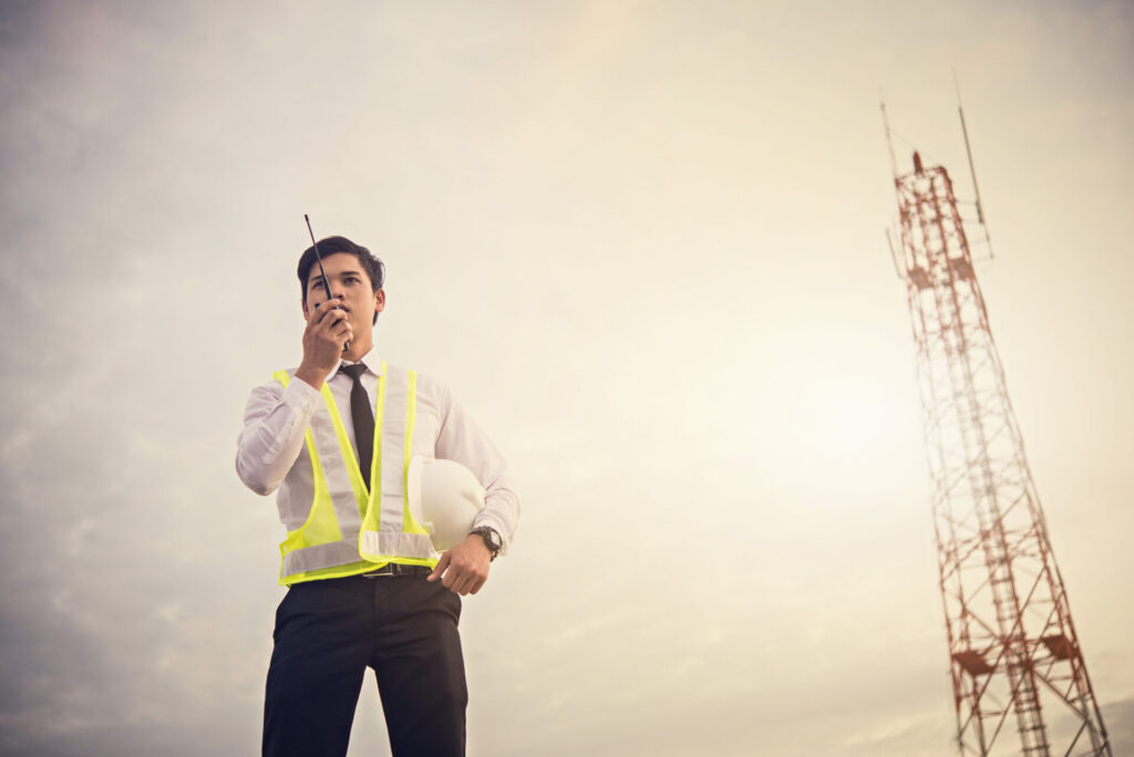 A male engineer working outdoors holding a helmet and using a radio to communicate.