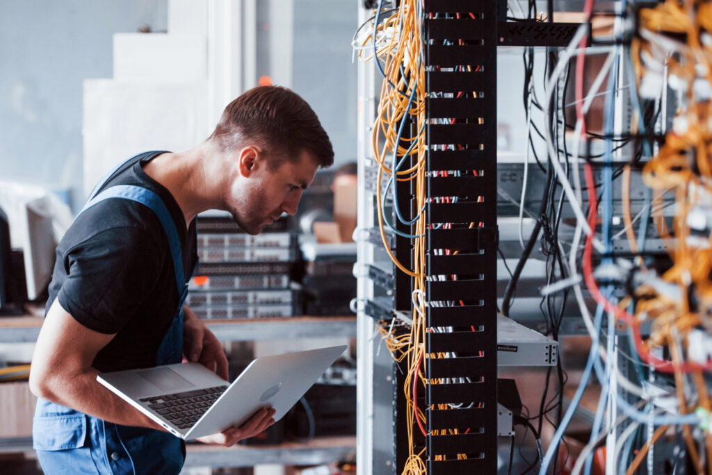 A male technician holding a laptop & working in a server room. 