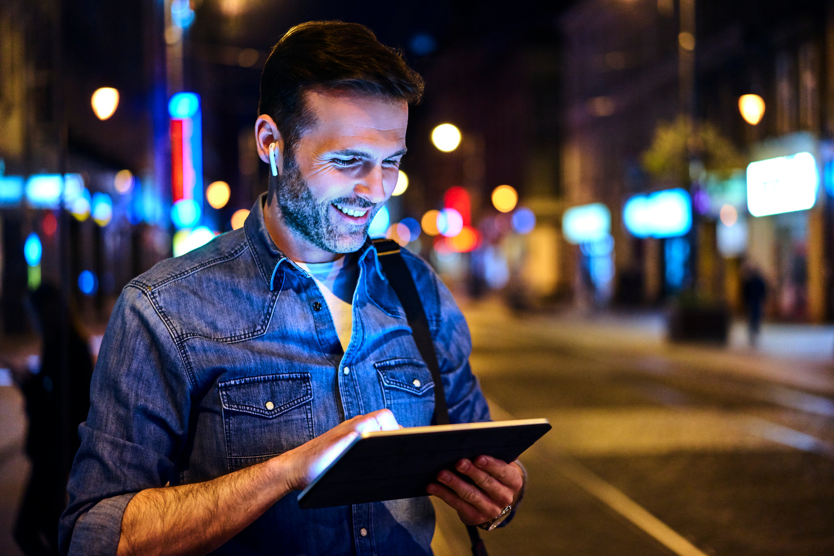 Portrait of man wearing wireless headphones and using tablet while standing at tram stop at night